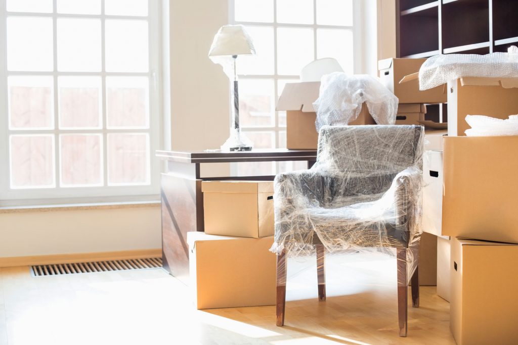 Moving boxes surround a bubble-wrapped chair and a table with a lamp.