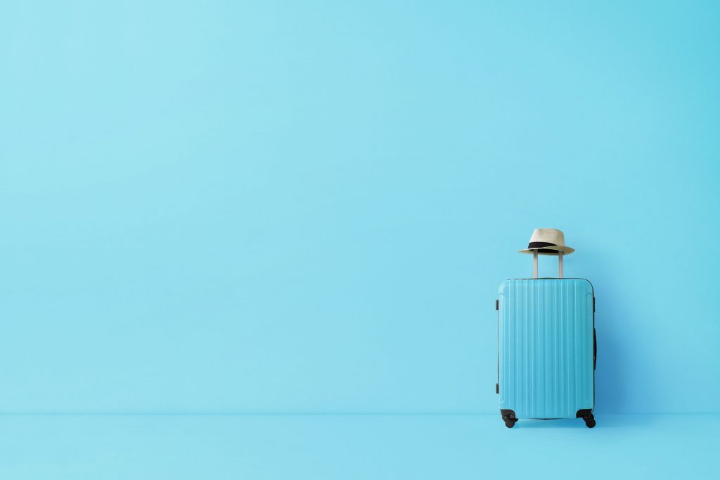 Blue, travelled-sized suitcase with a beige fedora on the handle, against a matching blue wall.