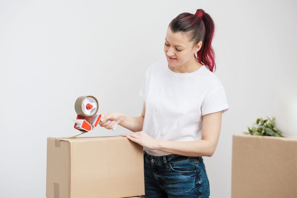 Person taping up a moving box with a tape roller, with another moving box waiting in the background.