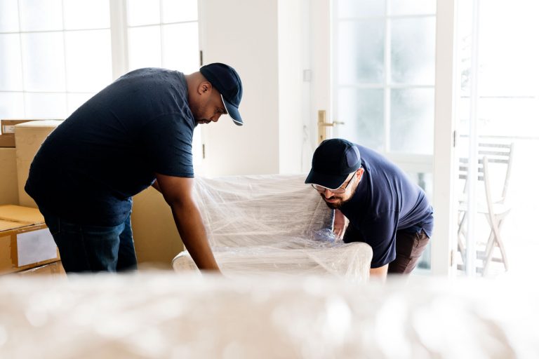 Two movers carrying a chair wrapped in bubble wrap. Other boxes and furniture are around them, waiting to be moved.